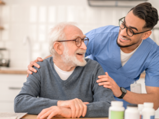 Consistency in Care shown as an older adult with white hair and glasses seated at a table, while a healthcare professional in blue medical scrubs stands nearby with a hand resting on the seated person’s shoulder; several medication bottles are arranged on the table in a bright kitchen setting.