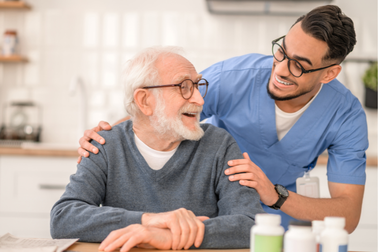 Consistency in Care shown as an older adult with white hair and glasses seated at a table, while a healthcare professional in blue medical scrubs stands nearby with a hand resting on the seated person’s shoulder; several medication bottles are arranged on the table in a bright kitchen setting.