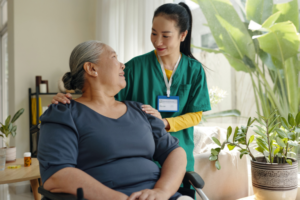 Older adult seated near a window holding a walking aid, receiving domiciliary care from another older adult standing beside them and placing a hand on the seated person’s shoulder in a softly lit home setting.