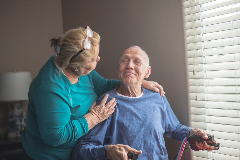 Older adult seated near a window holding a walking aid, with another older adult standing beside them and placing a hand on the seated person’s shoulder inside a softly lit room.